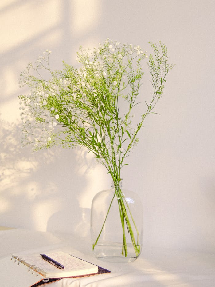 Artistic display of white wildflowers in a glass vase beside an open notebook in soft natural light.